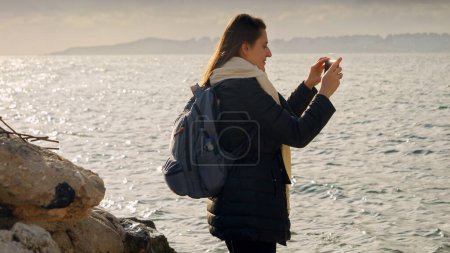 Photo for Young woman stands on the shoreline, capturing the winter ocean with her smartphone. Perfect for travel and vacation stock footage - Royalty Free Image