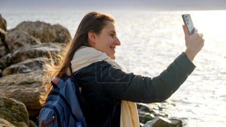 Foto de Mujer sonriente caminando y tomando fotos del mar tormentoso de invierno al atardecer usando su teléfono inteligente mientras está de pie sobre las rocas. Perfecto para viajes y turismo. - Imagen libre de derechos