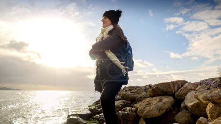 Photo for Smiling female tourist wit backpack wearing scarf and hat looking on cold sea waves and sunset. - Royalty Free Image