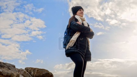 Foto de Un excursionista con una mochila de pie en un afloramiento rocoso en la cima de una montaña, contemplando la impresionante puesta de sol. Concepto de aventura al aire libre, exploración, viajes y naturaleza - Imagen libre de derechos