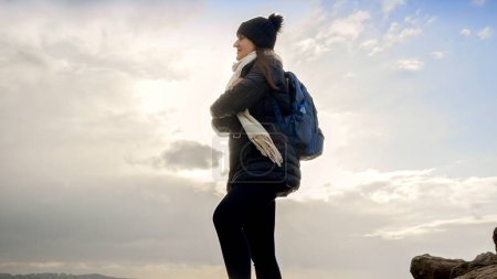 Téléchargez les photos : Jeune femme avec un sac à dos, profitant de la vue magnifique sur le coucher de soleil d'hiver sur la mer, du haut d'une falaise. Un concept d'aventure, de voyage et d'expérience de nouvelles choses - en image libre de droit