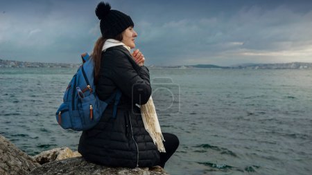 Photo for A woman with a backpack sitting on a cliff, taking in the beauty of the sunset over the cold, windy sea. A concept of adventure, travel, journey, and finding inner peace in nature - Royalty Free Image