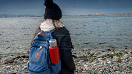 Foto de Vista trasera de la joven turista en ropa de invierno con mochila de pie en la playa de mar rocoso. - Imagen libre de derechos