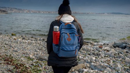 Photo for Young female hiker with backpack and thermos walking on the rocky sea beach at cold cloudy and windy day. Concept of hiking adventure, exploration, tourism and journey - Royalty Free Image