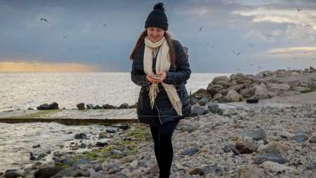 Téléchargez les photos : Femme souriante faisant une promenade sur la plage de l'océan par une journée nuageuse d'hiver et ramassant des coquillages. Concept de randonnée, voyage, exploration, tourisme et voyage - en image libre de droit