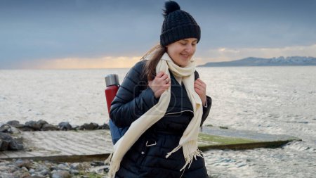 Téléchargez les photos : Une jeune femme avec un sac à dos et un thermos se promène le long d'une plage de mer rocheuse, souriant en admirant la vue imprenable. Parfait pour mettre en valeur la beauté de la nature et la joie de l'aventure - en image libre de droit