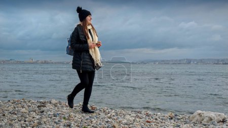 Foto de Mujer sonriente con sombrero y bufanda caminando por la playa del océano y recogiendo hermosas conchas marinas. Concepto de senderismo, viajes, exploración, turismo y viaje - Imagen libre de derechos