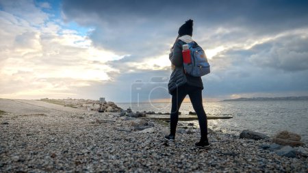 Photo for Young woman with a backpack and thermos takes a walk on a rocky sea beach, smiling as she explores the rugged terrain. Ideal for showcasing the journey and outdoor adventure - Royalty Free Image