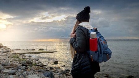 Foto de Caminante femenina en ropa de abrigo con una mochila y termo, caminando en una playa de mar rocoso en clima frío y ventoso. Perfecto para viajes, turismo y proyectos de aventura - Imagen libre de derechos