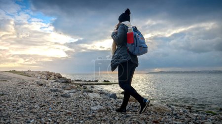 Foto de Senderista con ropa de abrigo equipada con una mochila y termo, disfrutando de un paseo por una playa rocosa de mar. Una adición perfecta a cualquier proyecto de viaje o turismo - Imagen libre de derechos