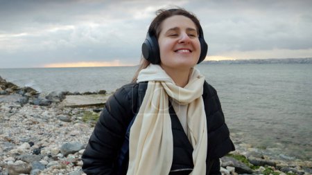 Téléchargez les photos : Femme souriante avec sac à dos marchant sur la plage de la mer par temps froid et venteux et écoutant de la musique dans les écouteurs. Concept de voyage, voyage, profiter de la musique - en image libre de droit