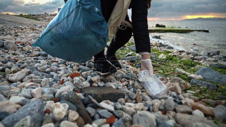 Foto de Primer plano de voluntaria recogiendo basura plástica en la playa del mar rocoso. - Imagen libre de derechos