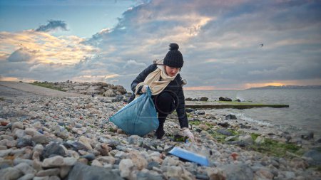 Téléchargez les photos : Une jeune femme bénévole en action, portant des gants et ramassant des ordures sur le rivage, soulignant l'importance de maintenir des océans propres et sains - en image libre de droit