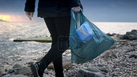 Téléchargez les photos : Femme bénévole marchant avec un sac poubelle rempli de déchets sur la plage de la mer rocheuse. - en image libre de droit