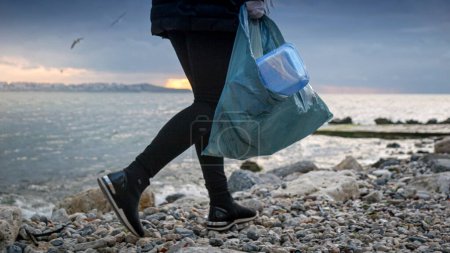 Foto de Mujer voluntaria en acción, caminando por la playa del mar y recogiendo residuos, mostrando sus esfuerzos para proteger el medio ambiente y promover hábitos ecológicos - Imagen libre de derechos