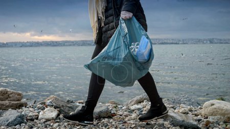 Photo for Closeup of woman volunteer holding plastic bag full of garbage and trash after cleaning rocky sea beach. - Royalty Free Image