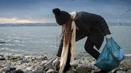 Téléchargez les photos : Mouvement lent d'une femme en gants de caoutchouc marchant sur la plage, ramassant les déchets plastiques et les débris, et les mettant dans un sac poubelle. Un concept d'écoconscience, d'environnementalisme et de conservation - en image libre de droit