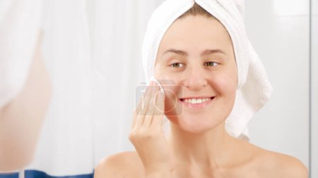 Téléchargez les photos : Femme souriante en serviette de bain nettoyage visage avec lotion après avoir pris une douche. Concept de belle femme, maquillage à la maison, soins de la peau et industrie de la beauté domestique - en image libre de droit