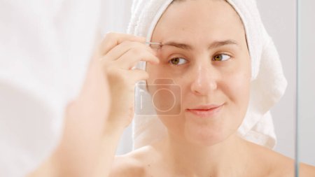 Foto de Mujer joven sonriente con una toalla blanca arrancándose las cejas con pinzas cosméticas. Concepto de hermosa hembra, maquillaje en el hogar, cuidado de la piel y la industria de la belleza doméstica - Imagen libre de derechos