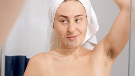 Téléchargez les photos : Jeune femme souriante levant le bras et regardant les longs poils des aisselles après avoir pris une douche. Concept de belle femme, beauté naturelle, féminité et poils du corps - en image libre de droit