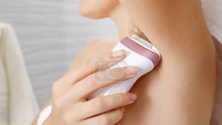 Photo for Young woman using electric epilator to remove armpit hair in bathroom. - Royalty Free Image