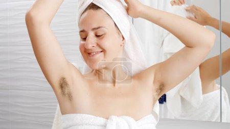 Téléchargez les photos : Belle femme torse nu en serviette de bain montrant ses longs cheveux aisselle au miroir de salle de bains. Concept d'hygiène, beauté naturelle, féminité et croissance des poils du corps - en image libre de droit