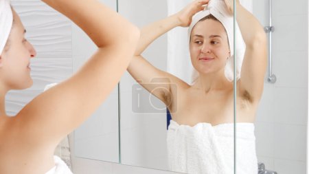 Téléchargez les photos : Femme souriante dans une serviette de bain soulevant les bras et montrant de longs cheveux foncés sur les aisselles. Concept d'hygiène, beauté naturelle, féminité et croissance des poils du corps - en image libre de droit