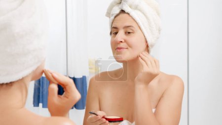 Téléchargez les photos : Belle jeune femme en serviette de bain appliquant de la poudre visage au miroir. Concept de belle femme, maquillage à la maison, soins de la peau et industrie de la beauté domestique. - en image libre de droit