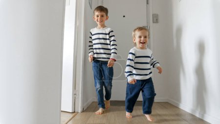 Photo for Two little boys running on a wooden floor towards their mother, who welcomes them with a warm hug. Love and happiness that can be found within a family home - Royalty Free Image