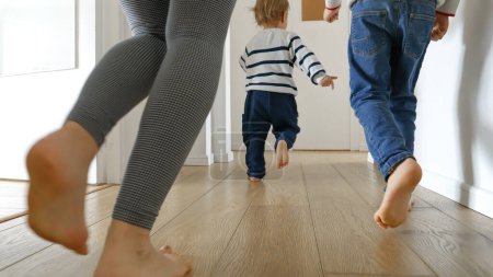 Photo for Closeup of mother and two boys running on the wooden floor at home. Concept of family love, joy, and fun in a household. - Royalty Free Image