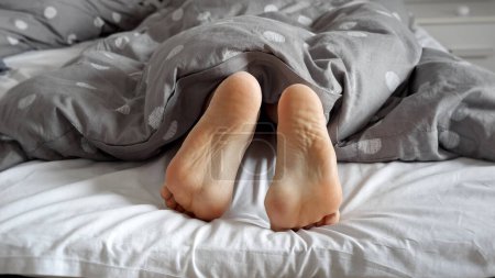 Téléchargez les photos : Gros plan de pieds féminins couchés sur le lit et montrant sous la couverture, femme se retournant et cherchant une pose confortable. Concept de détente à la maison, lit confortable, repos le matin - en image libre de droit