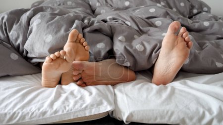 Téléchargez les photos : Gros plan des pieds du couple reposant sur un lit doux pendant qu'ils profitent d'un moment de calme ensemble. Concept d'amour, de convivialité et de détente à la maison - en image libre de droit