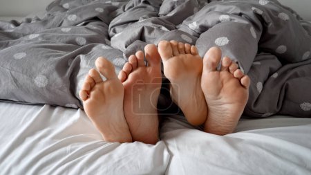 Téléchargez les photos : Gros plan comme les pieds d'un homme et d'une femme se trouvent enlacés sous la couverture sur un lit doux. Sentiments de chaleur, d'amour et d'affection dans un cadre familial confortable - en image libre de droit
