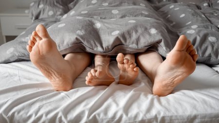 Téléchargez les photos : Gros plan des pieds aimants des parents et de l'enfant couchés sous la couverture sur le lit. Concept d'amour familial, d'enfance heureuse, de plaisir à la maison, de parentalité, de relation et de vie familiale - en image libre de droit