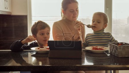 Téléchargez les photos : Deux petits garçons avec leur mère mangeant de la soupe assis sur la cuisine et regardant la vidéo sur tablette. - en image libre de droit