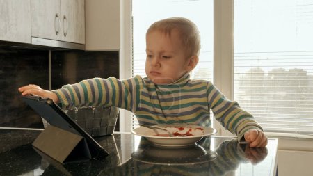 Téléchargez les photos : Petit garçon bouleversé regarder des dessins animés sur tablette tout en mangeant de la soupe sur la cuisine. Nourriture domestique, cuisine à la maison, alimentation saine des enfants - en image libre de droit