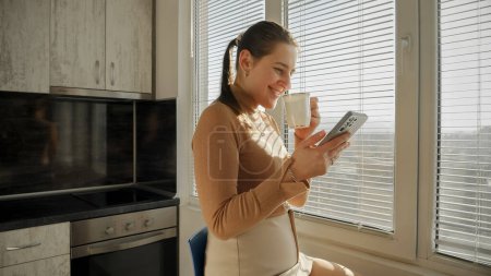 Téléchargez les photos : Femme souriante naviguant sur Internet sur smartphone et buvant du café le matin. Les gens le matin, beau paysage urbain, ayant une pause au bureau - en image libre de droit