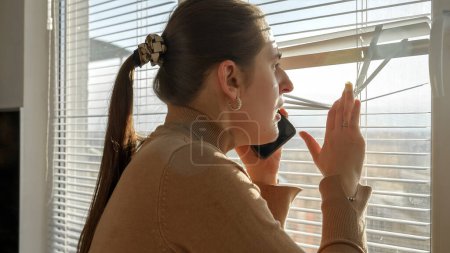 Photo for Young worried woman talking by phone and looking through window blinds. Crime witness, spying through window, peeking on street - Royalty Free Image