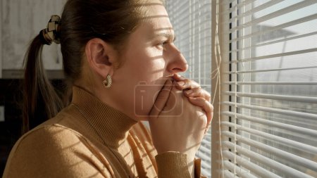 Ängstliche und verängstigte Frau schließt den Mund mit der Hand und schaut durch Jalousien aus dem Fenster.