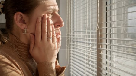 Foto de Mujer joven siendo testigo de algo aterrador o criminal mientras mira a través de persianas de ventana. - Imagen libre de derechos
