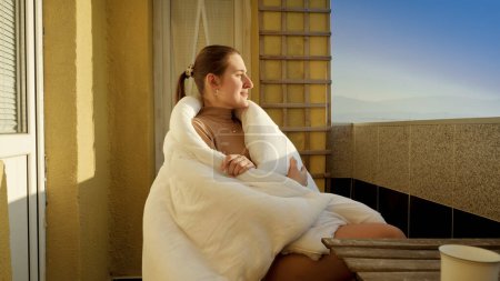 Photo for Young woman sitting on terrace in blanket and looking at sun rays. People in morning, relaxing at home, enjoying city view - Royalty Free Image