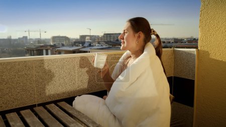 Téléchargez les photos : Joyeux sourire femme boire du café sur le balcon et profiter du soleil du matin. Les gens le matin, se détendre à la maison, profiter de la vue sur la ville - en image libre de droit
