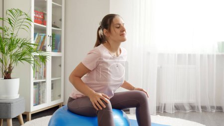 Téléchargez les photos : Belle jeune femme assise sur fitball et faisant des exercices de fitness à la maison. Concept de santé, de sport et de yoga à la maison. - en image libre de droit