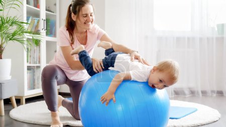 Photo for Smiling mother doing stretching exercises to her baby son rollling on fitness ball at home. - Royalty Free Image