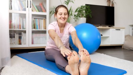 Téléchargez les photos : Femme souriante heureuse en leggings étirant et atteignant ses orteils tout en faisant de l'exercice sur tapis de fitness. Concept de santé, de sport et de yoga à la maison - en image libre de droit