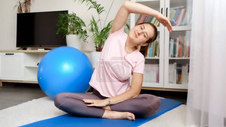 Photo for Young relaxed woman sitting on yoga mat at living room and stretching hand and back muscles. - Royalty Free Image