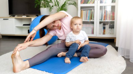 Photo for Cute baby boy sitting on fitness mat and looking at his mother stretching legs before sports training. Concept of healthcare, sports and yoga at home - Royalty Free Image