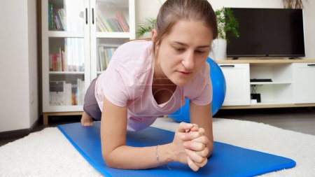 Téléchargez les photos : Portrait de jeune femme concentrée debout en position de planche sur tapis de fitness dans le salon. Concept de santé, de sport et de yoga à la maison - en image libre de droit
