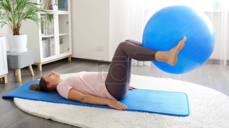 Photo for Young brunette woman lying on fitness mat and lifting big fit ball with legs. - Royalty Free Image