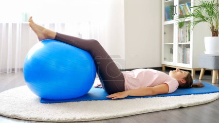 Foto de Hermosa mujer joven acostada en la alfombra de fitness y el entrenamiento con fitball azul. Concepto de salud, deportes y yoga en casa - Imagen libre de derechos